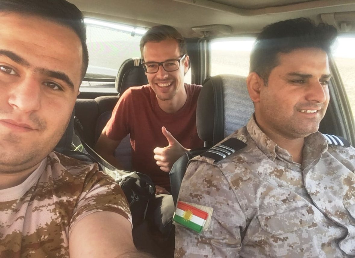 Ruben Vermeer in the back of a car between two Kurdish security men in camouflage, one with a Kurdistan flag patch, giving a thumbs-up