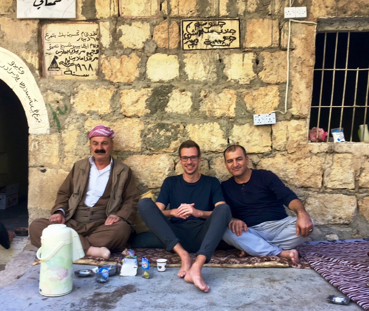 Ruben Vermeer sharing tea on carpets with two Yazidi men against the old stone wall of the sanctuary complex at Lalish
