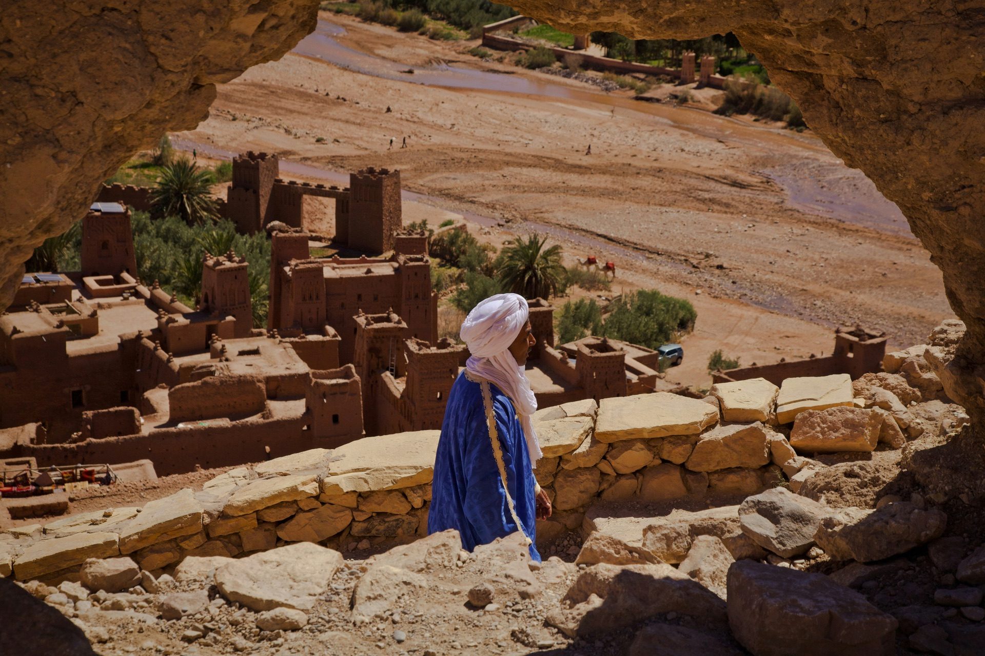 An Amazigh man in indigo dress leading a camel across desert terrain