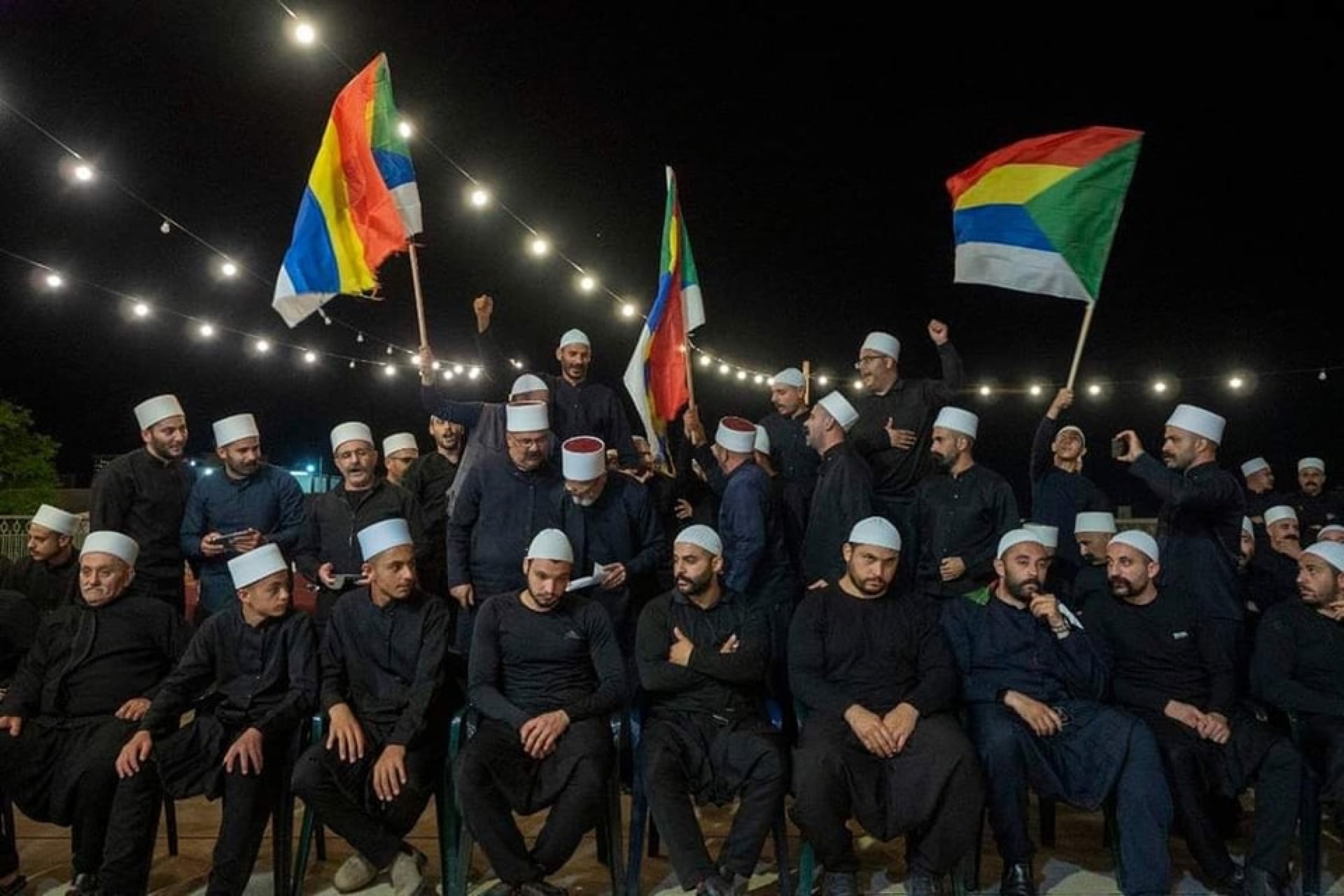 Druze men in traditional white caps gathered at night under festival lights, waving the five-coloured Druze flag
