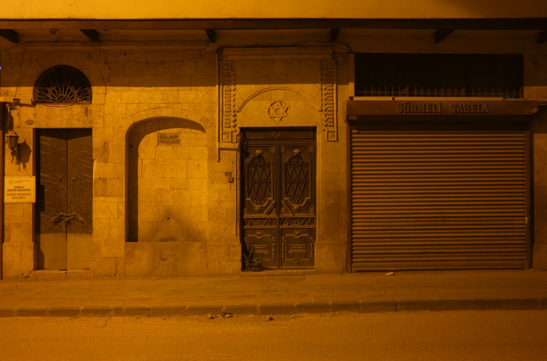 The shuttered stone facade of the synagogue of Antakya at night, warmly lit from the street