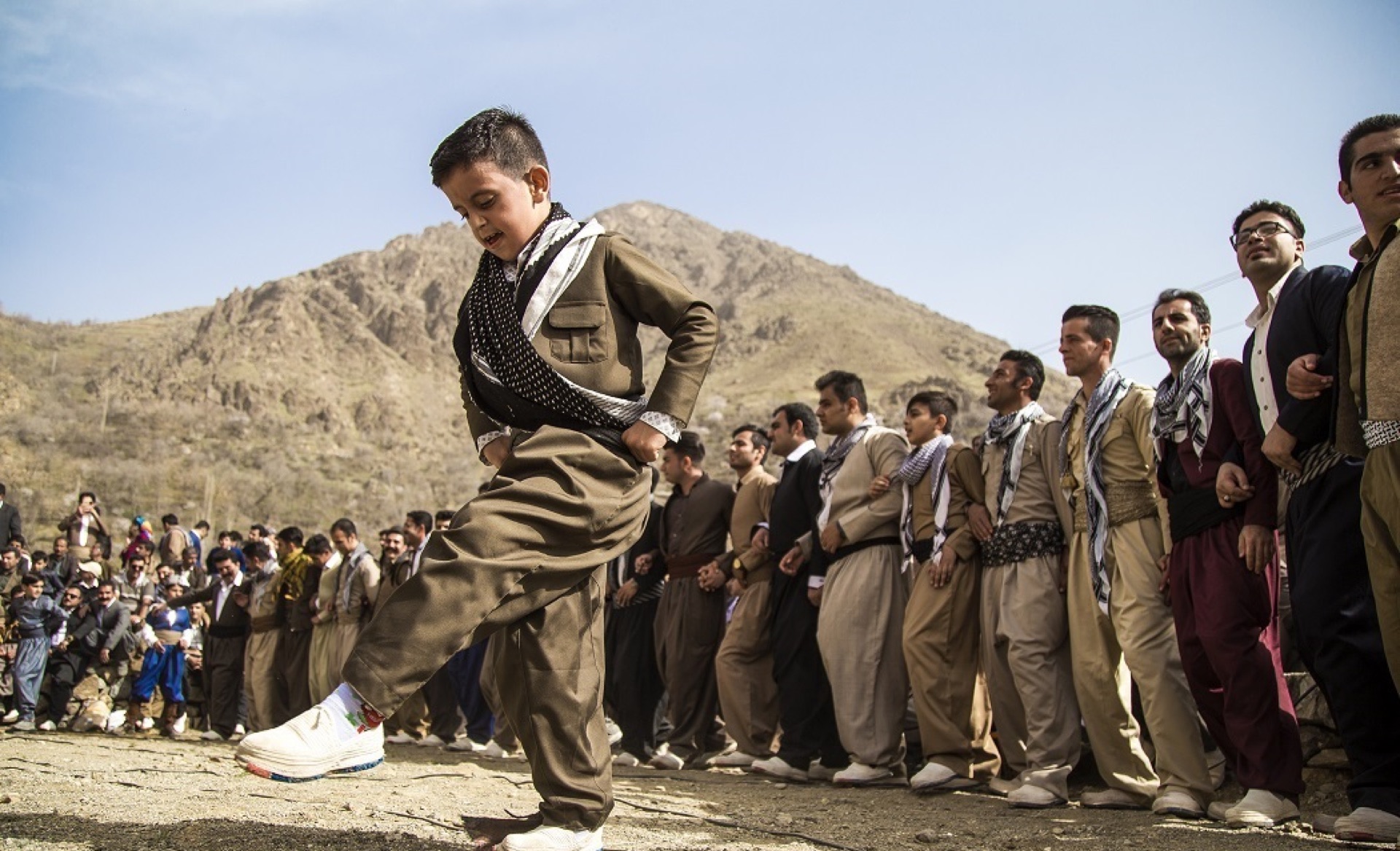 A boy in traditional Kurdish dress dancing before a long line of men at a mountain Newroz celebration