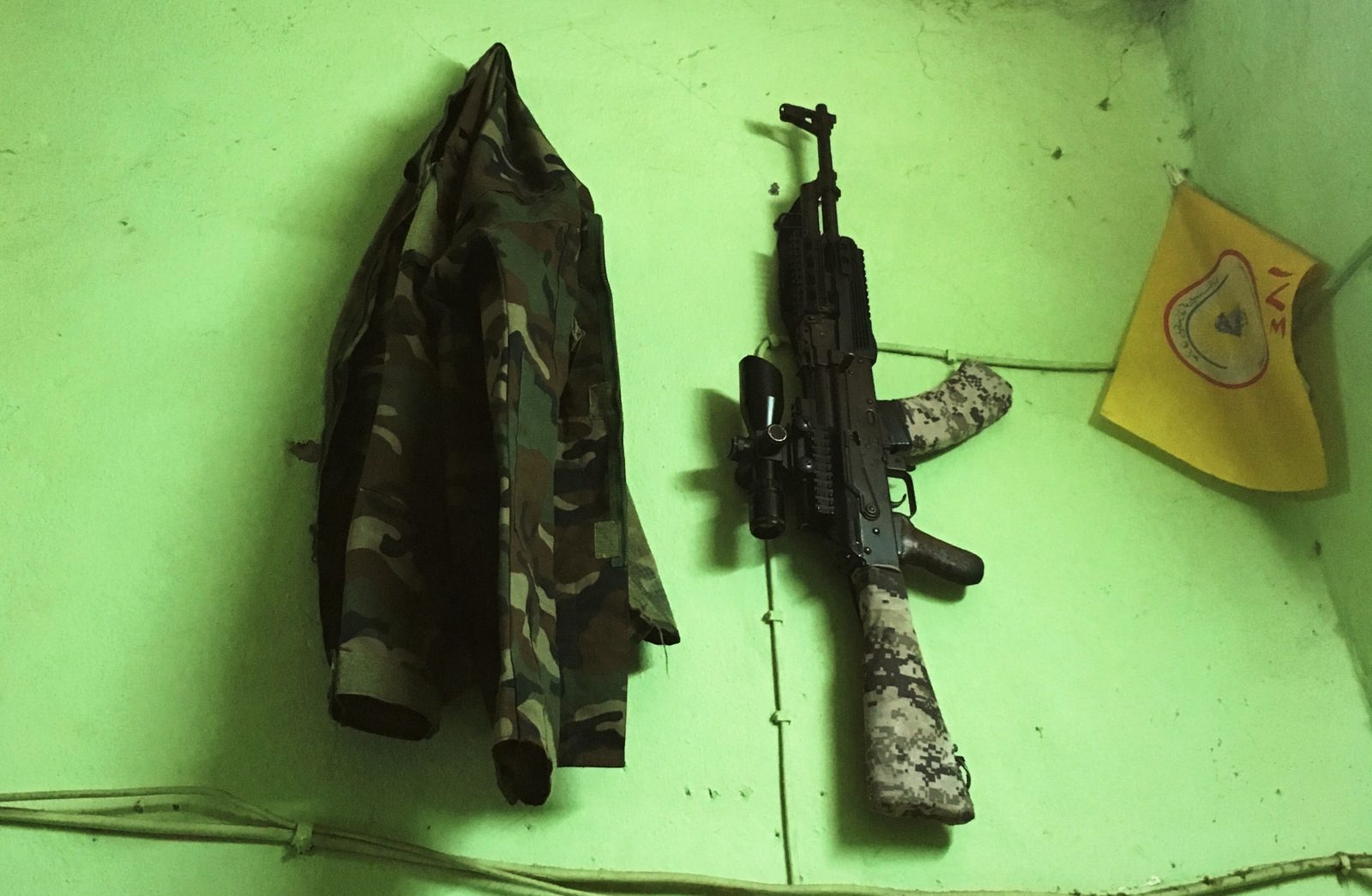 A camouflage jacket and a Kalashnikov hanging side by side on a lime-green barrack wall