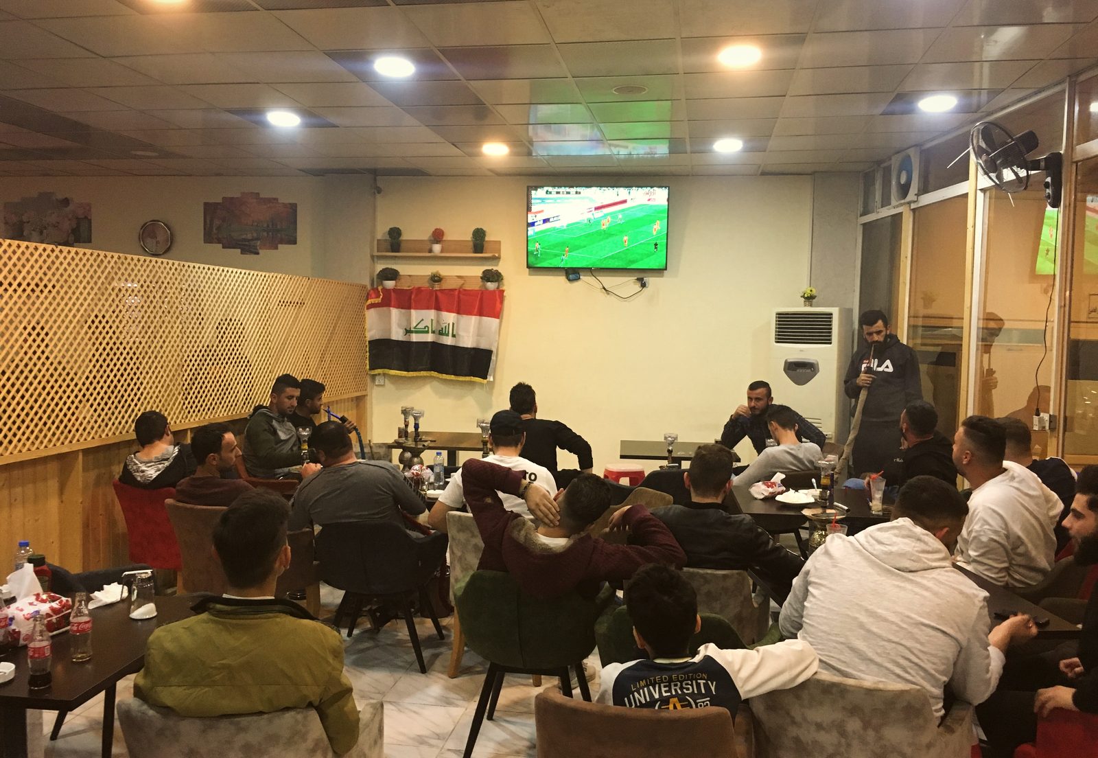 A café full of young men watching Iraq play Oman on a wall-mounted television, an Iraqi flag hanging beside the screen
