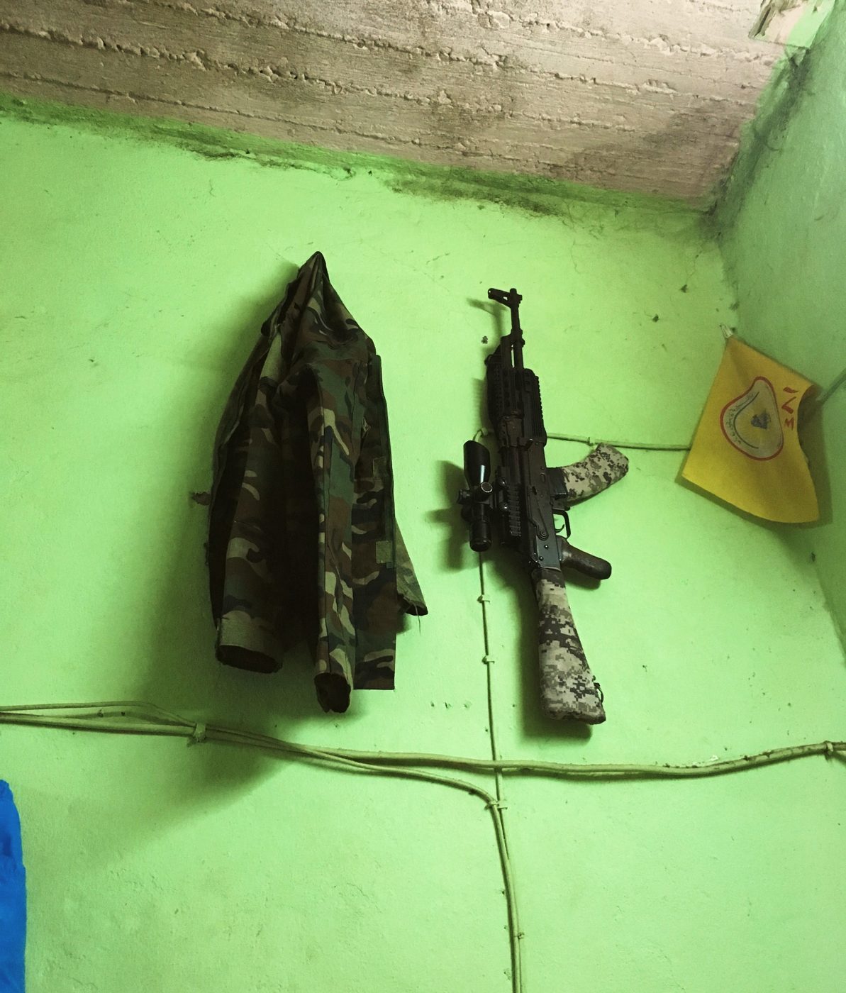 A camouflage jacket and a Kalashnikov hanging on the lime-green wall of the barrack