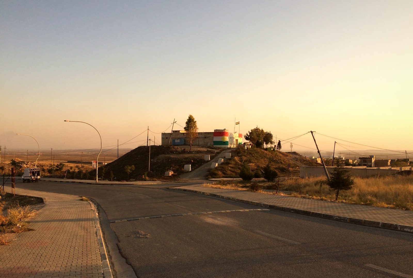 An empty road at dusk outside Alqosh, leading toward a low hill with buildings and flags