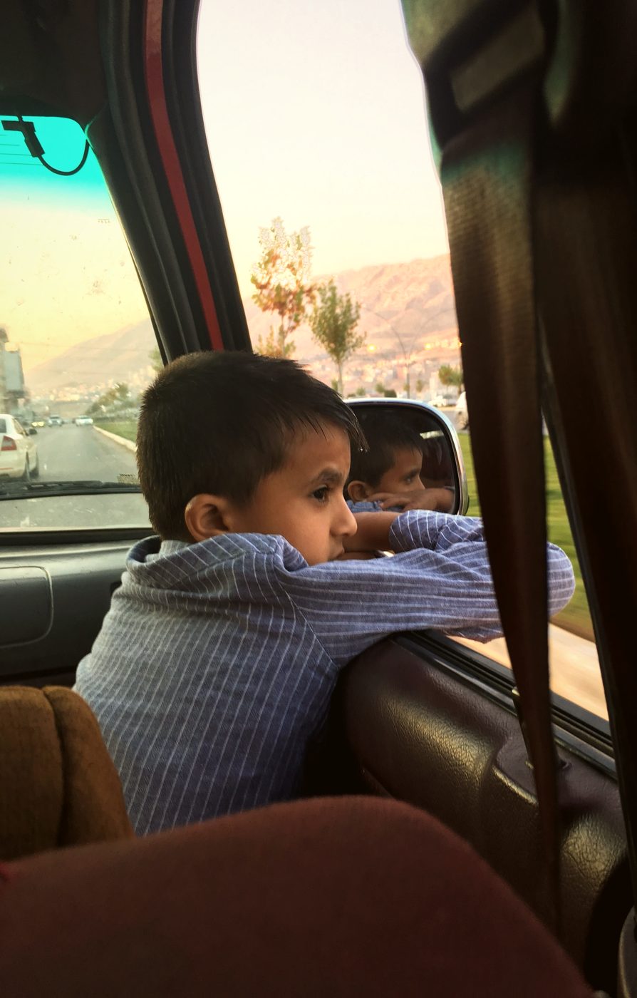 A young boy leaning on a car window ledge at dusk, gazing out at hills and a distant town