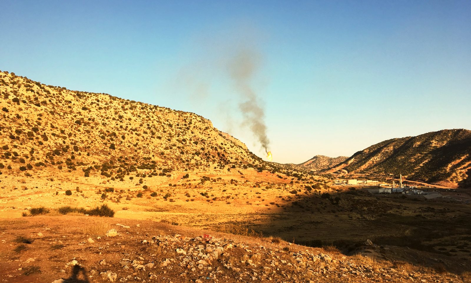 Golden scrub hills in northern Iraq at low sun, a plume of smoke rising from a gas flare beyond the ridge