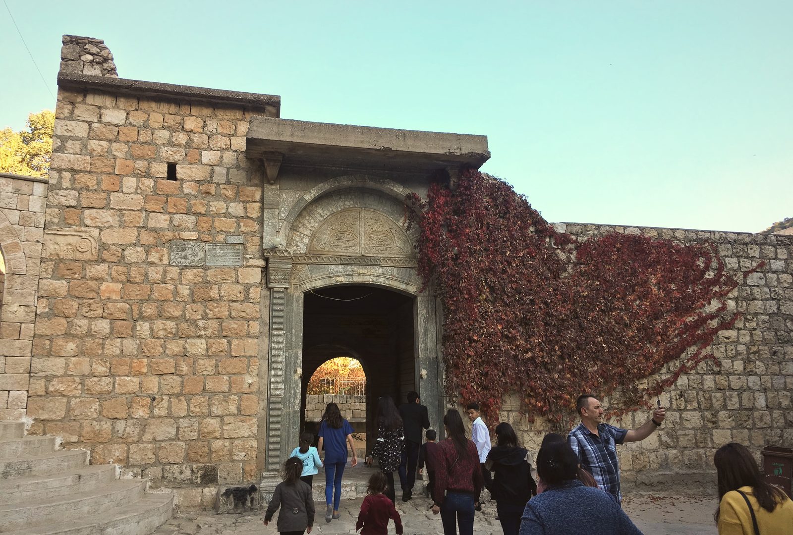 The carved stone gateway of Lalish under scarlet autumn ivy, pilgrims walking up the steps into the arch