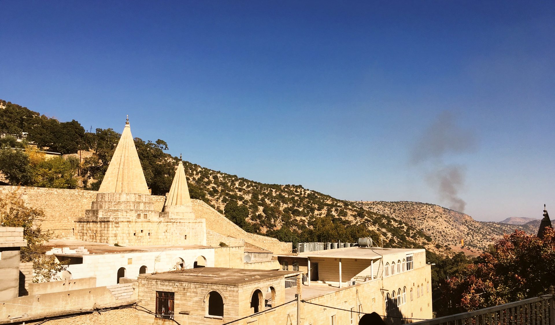 The conical spires of Lalish rising above the sanctuary rooftops, with a smoke plume over the hills beyond