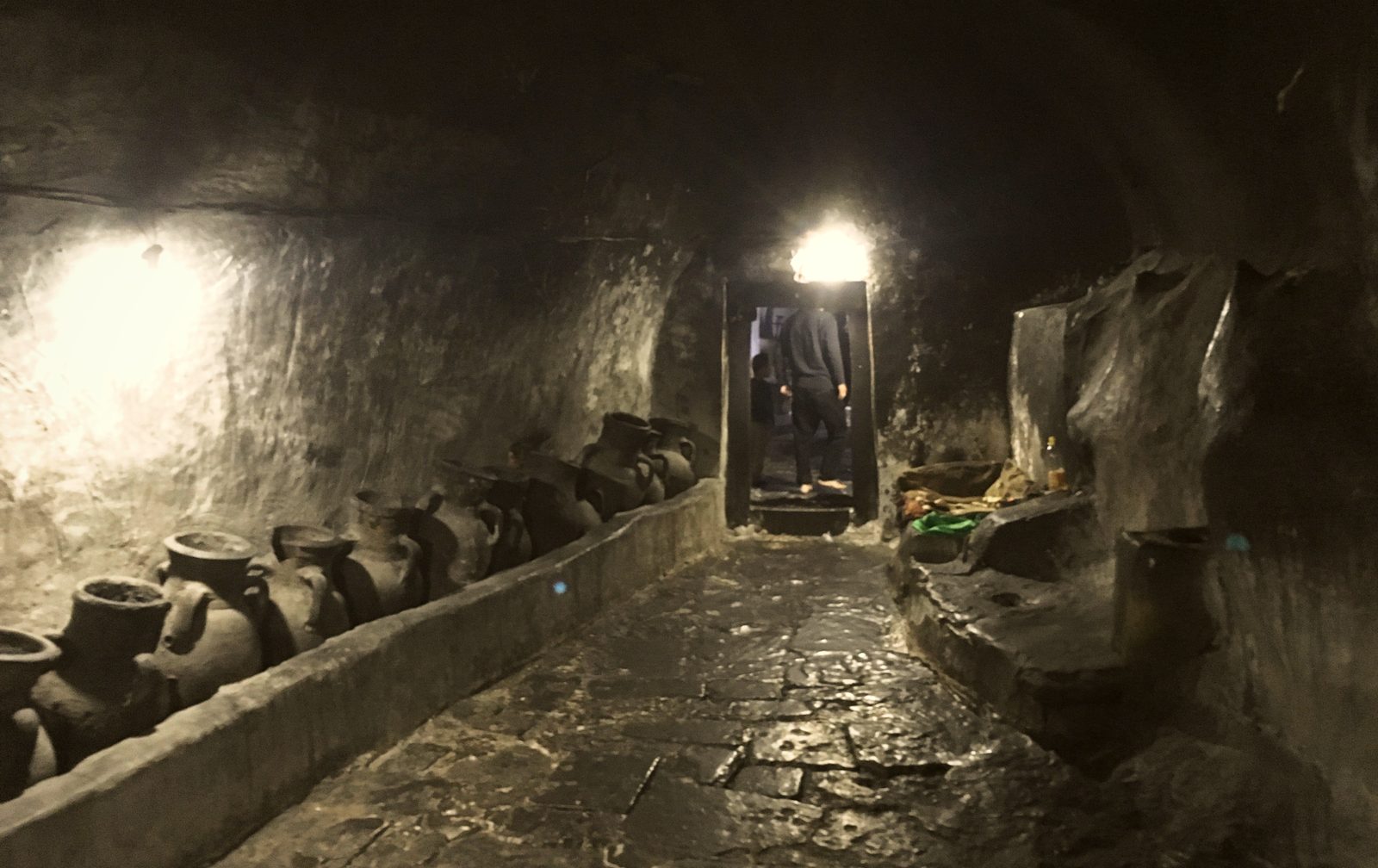 A dark vaulted tunnel inside the Lalish temple, rows of earthenware oil jars along the wall and a lit doorway beyond