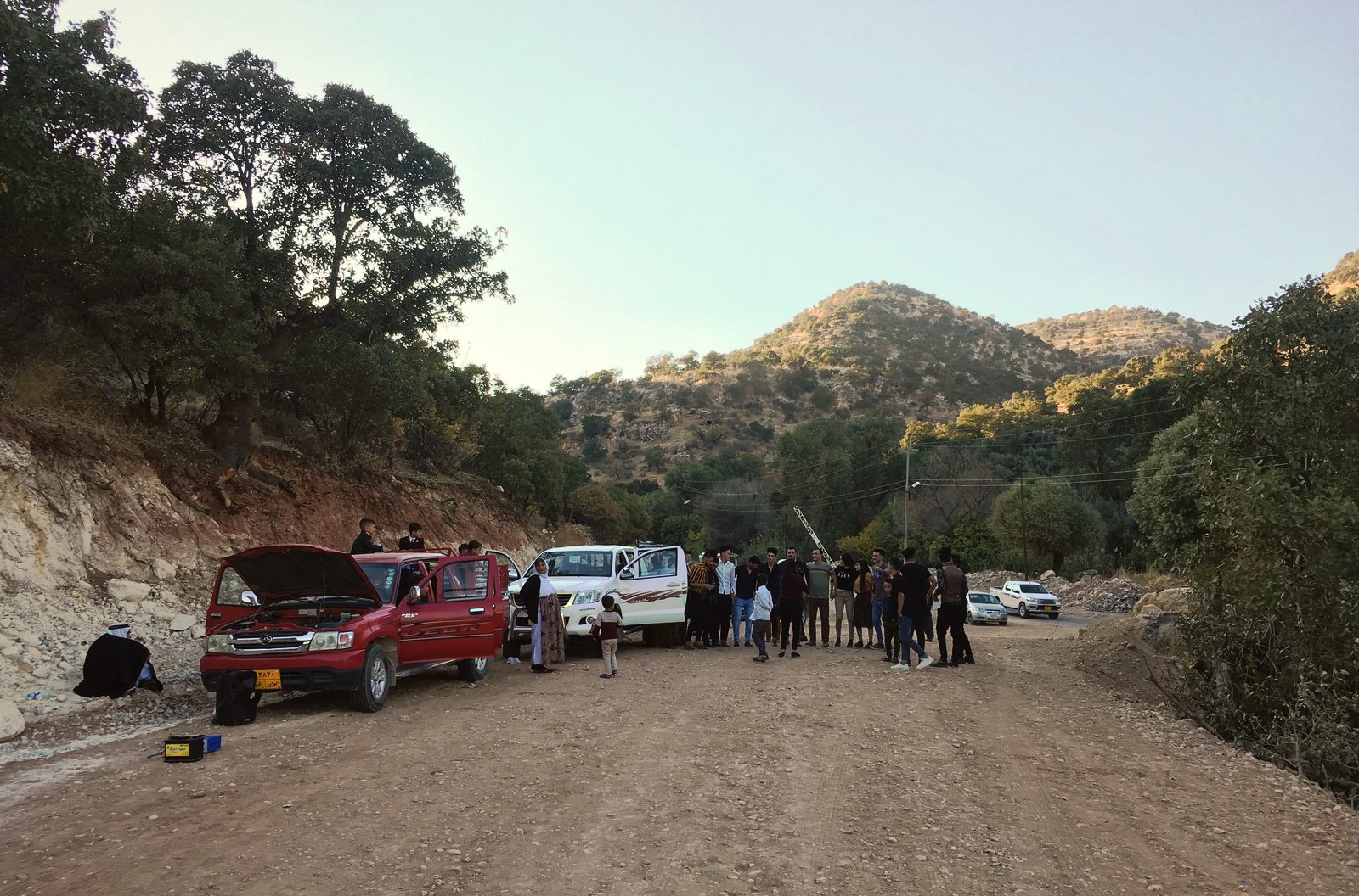 A dirt road outside Dohuk with parked cars and a group of people standing in the afternoon light