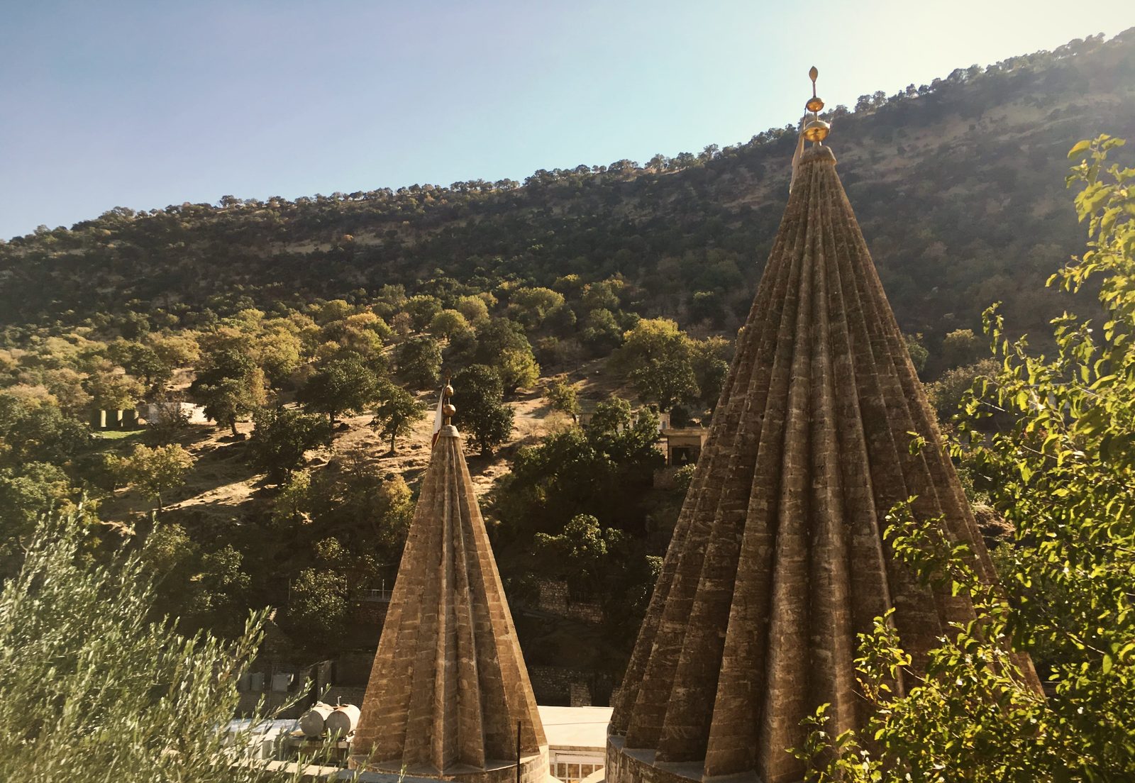 Two fluted conical spires of the Lalish sanctuary, one topped with a gilded finial, against a wooded hillside