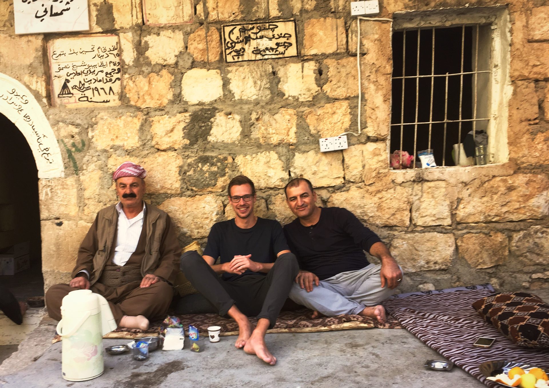 The author sitting barefoot on rugs with two Yazidi men outside a stone building, tea things spread between them