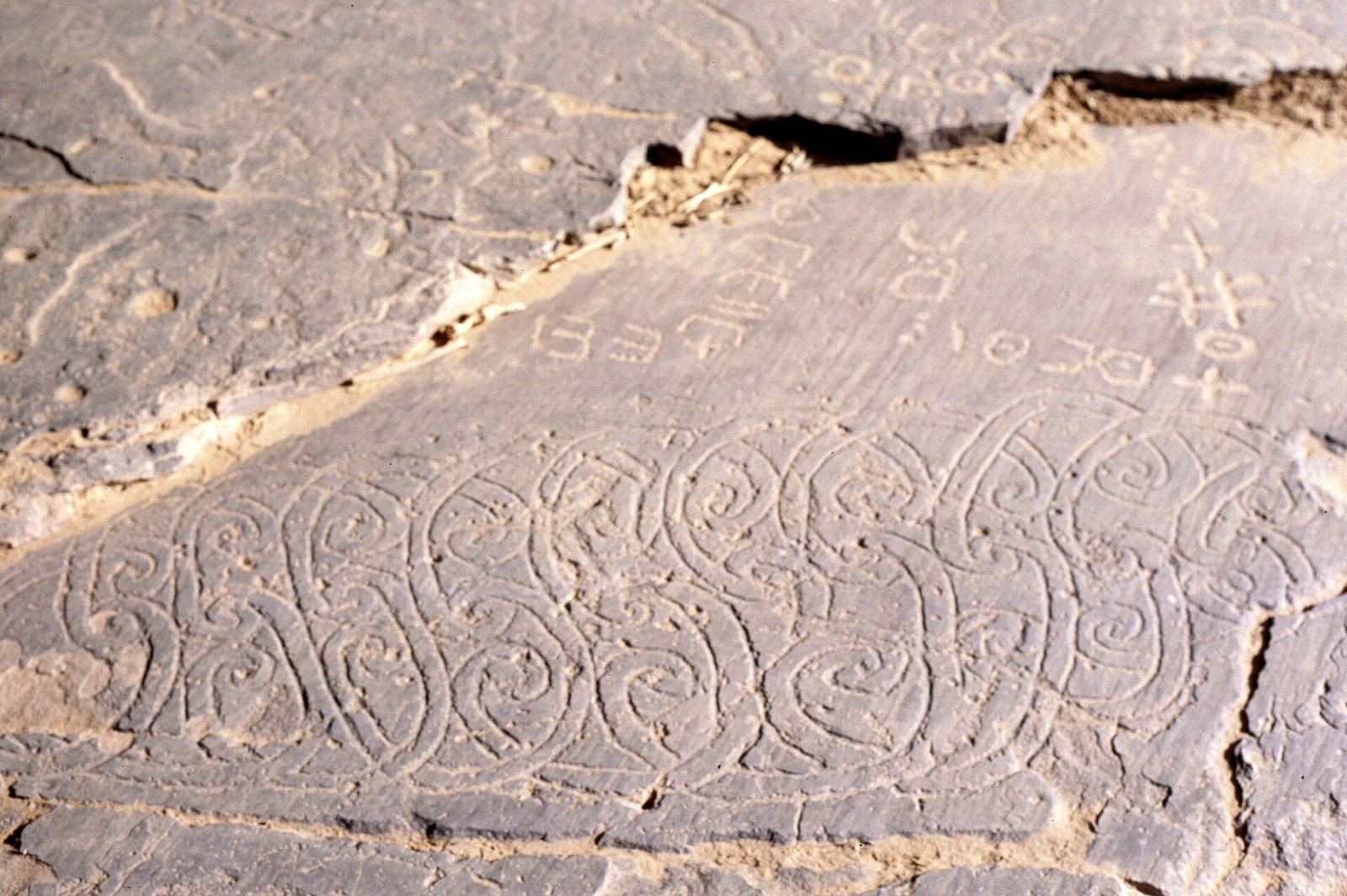 Angular Tifinagh characters and spiral engravings carved into a pale rock surface in the Algerian Sahara